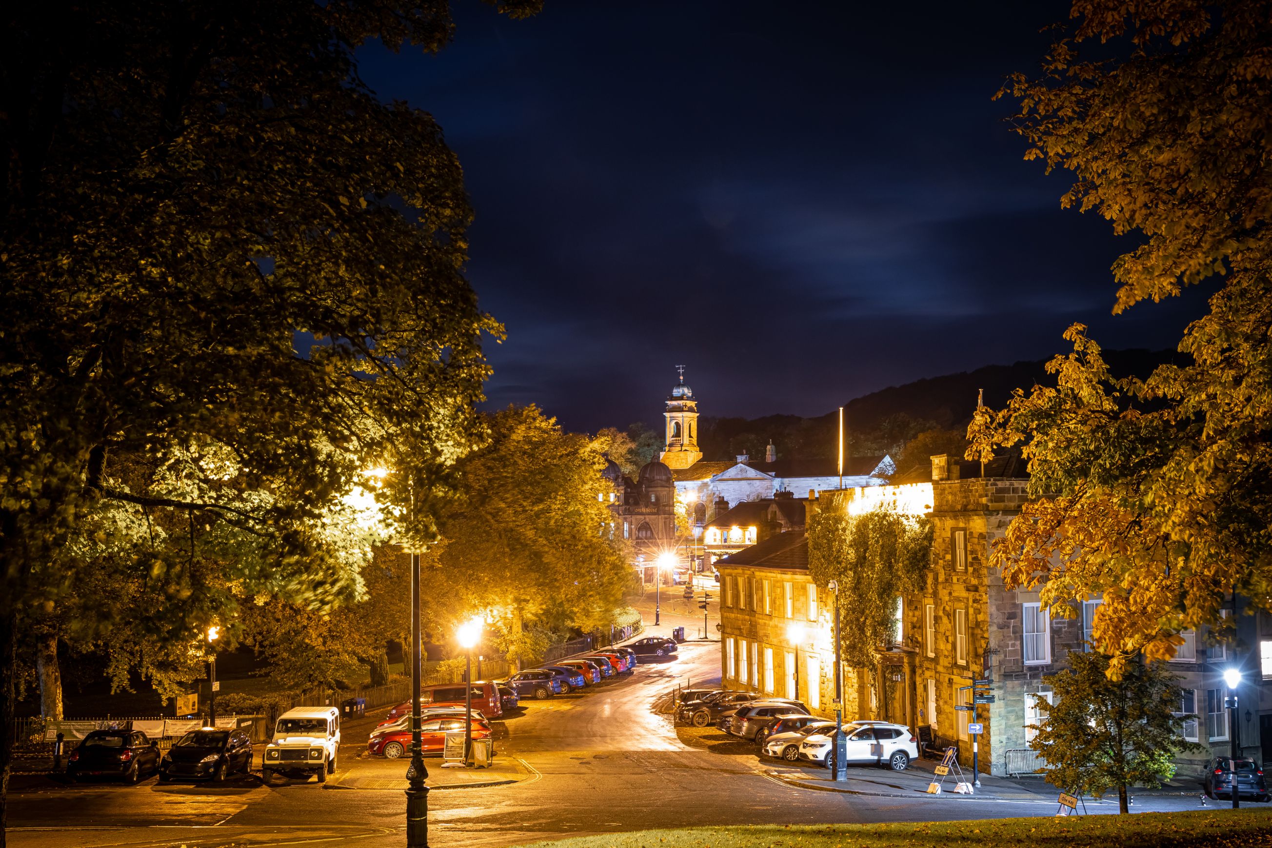 Shot of a road in the town of Buxton at night time, showing trees, houses, parked cars and St John's Church