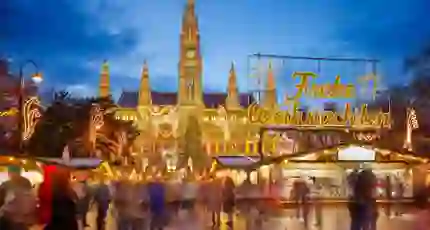 Shot of Vienna's gothic city hall which has one tall clock tower and four smaller towers, two on each side, all with pointy turrets. The building is a gold colour and is in front of a bright blue night sky. In front, is a Christmas Market, showing strips of cabin-like stalls and a variety of lit up Christmas decorations, one in the forefront reading 'Merry Christmas' in Austrian. People can be seen, although blurry as they are moving around.