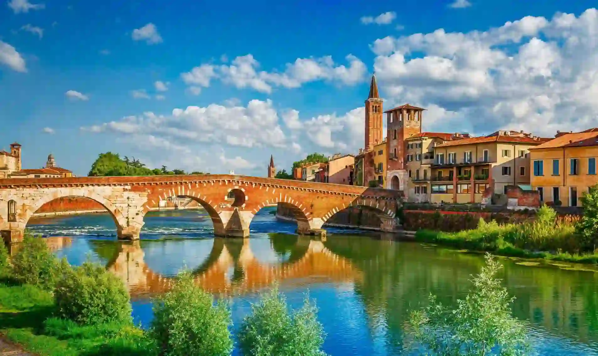 Scenic view of the historic Ponte Pietra bridge crossing the Adige River in Verona, Italy, with colourful old buildings and church towers under a bright blue sky with scattered clouds