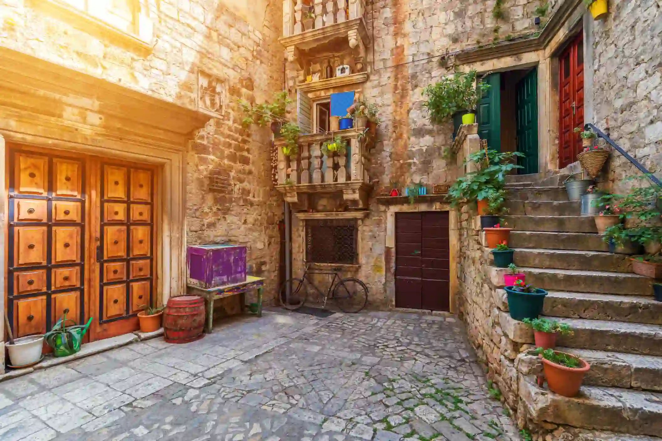 Shot of a concrete yard with steps on the right, with flower pots going up them, leading to a green door and a blue door. in the centre, a bicycle is parked up a stone wall, below two small Juliette balconies, then to the left there is a wide wooden door in a stone wall, and below it, empty flower pots, a table and a box.