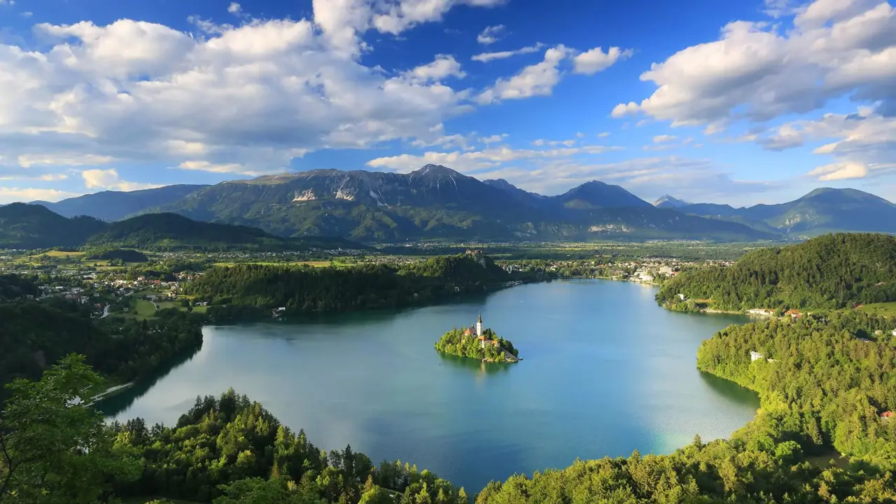 Wide angle shot of Lake Bled, showing its forested surrounding areas, and mountains in the distance. In the middle of the lake, there is a small island with trees and a towered building
