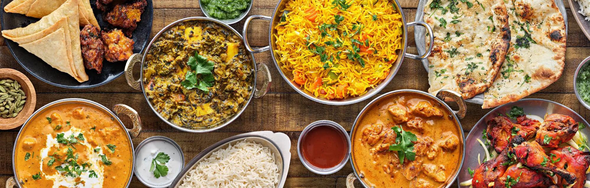 A colourful spread of Indian food featuring dishes such as curry, naan bread, rice, samosas, and chutneys served in bowls and plates on a wooden table