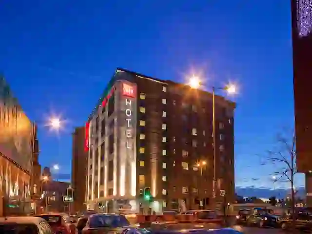 Exterior view of the ibis Belfast City Centre hotel at dusk, with the building lit up and traffic passing by