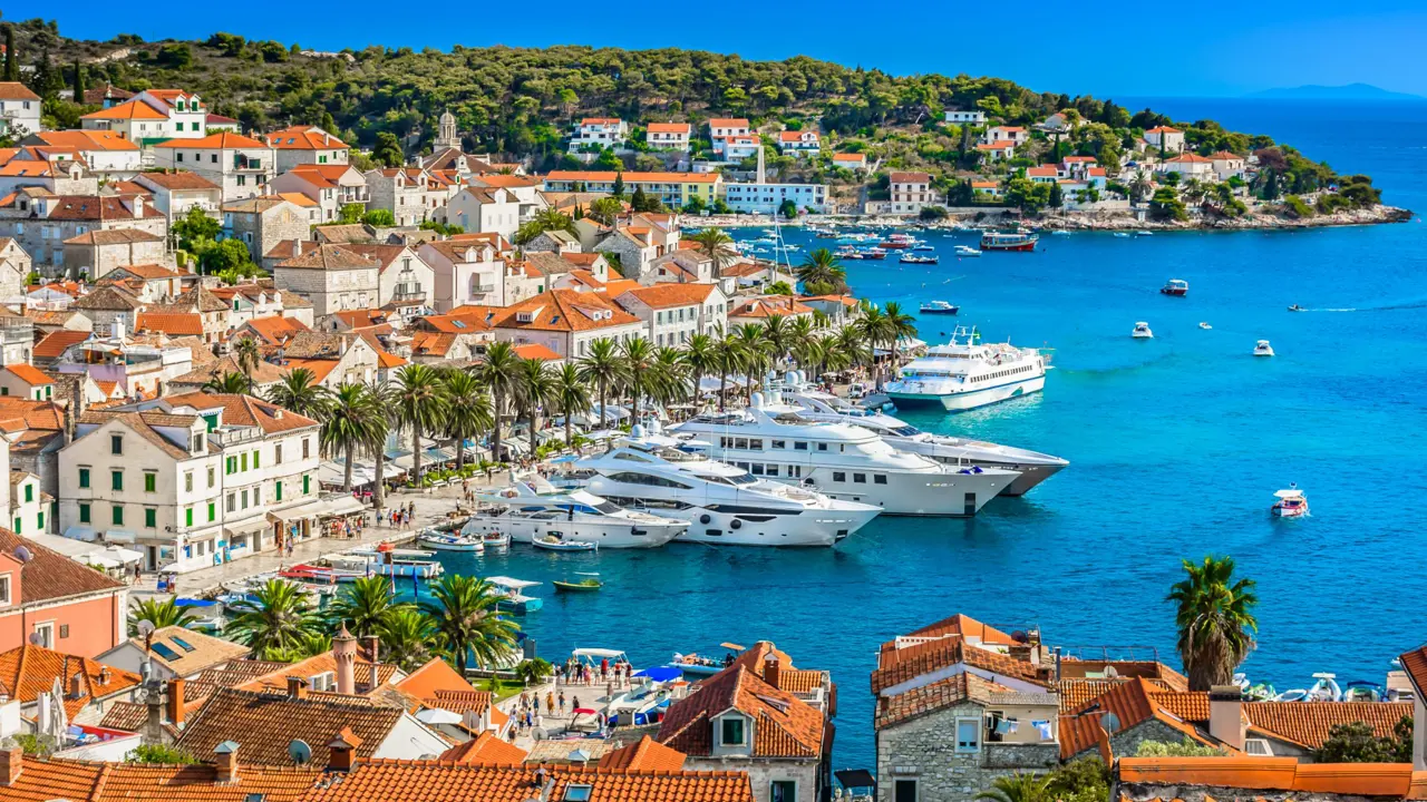 High angle shot of a town on the waterfront, with yachts docked on the water. Rows of palm trees all round the water's edge, in front of cream houses with orange roofs, and orange roofs in the forefront. Further away, land covered in trees and a blue sky.