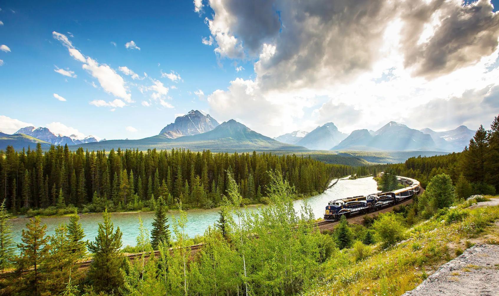 The Rocky Mountaineer train travelling through the Canadian Rockies, with rugged mountains in the background and dense evergreen forests lining the tracks