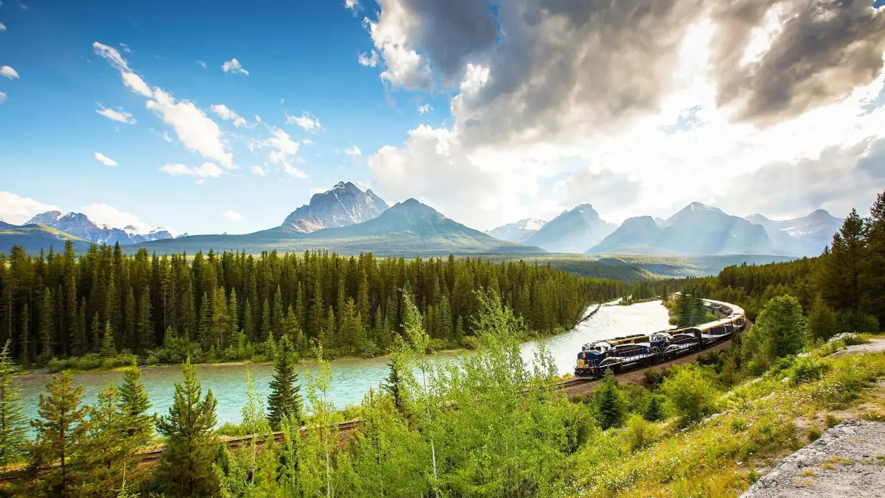 The Rocky Mountaineer train travelling through the Canadian Rockies, with rugged mountains in the background and dense evergreen forests lining the tracks